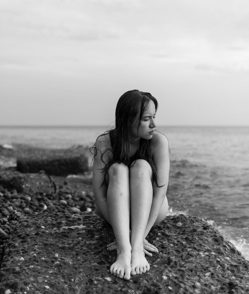young naked woman sitting curled up on stony beach