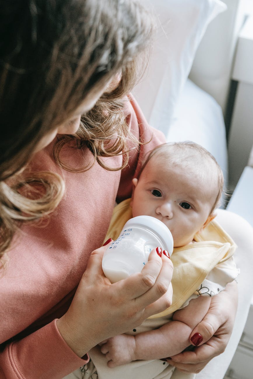 cute newborn drinking milk from bottle in hands of crop mom