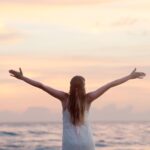 rear view of woman with arms raised at beach during sunset